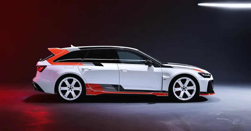 Side view of a white Audi RS6 Avant GT station wagon with orange and black stripes, white rims, and a blacked-out roof, in a studio setting with a dark background and soft lighting.