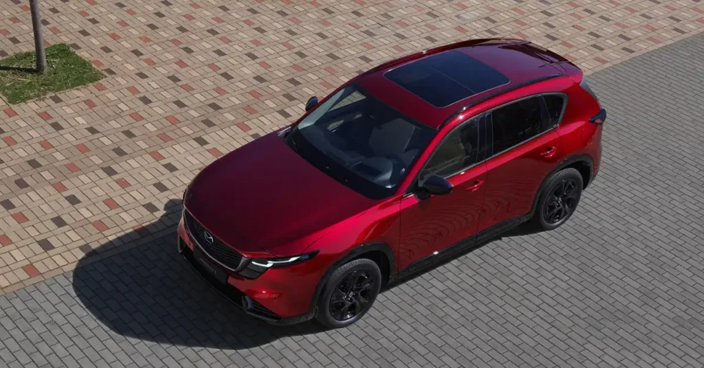 An eye-level shot captures a red Mazda CX-50 SUV parked on a paved surface, with a small patch of grass and a tree trunk visible in the upper left corner.
