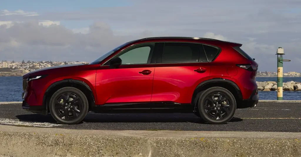 A side view of a red Mazda CX-5 parked on a concrete surface near a body of water, with a city skyline visible in the background.