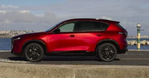 A side view of a red Mazda CX-5 parked on a concrete surface near a body of water, with a city skyline visible in the background.