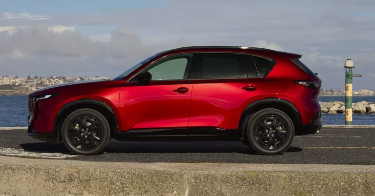 A side view of a red Mazda CX-5 parked on a concrete surface near a body of water, with a city skyline visible in the background.