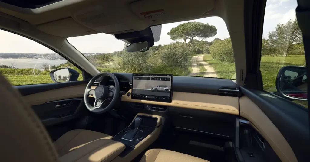Interior of a Mazda CX-50 SUV, with a black dashboard and a beige trim, a steering wheel with the Mazda logo, a large touchscreen display showing a car, a gear shift, and seats, with a view of a dirt road, green grass, trees, and a body of water in the background, captured with a wide-angle lens.
