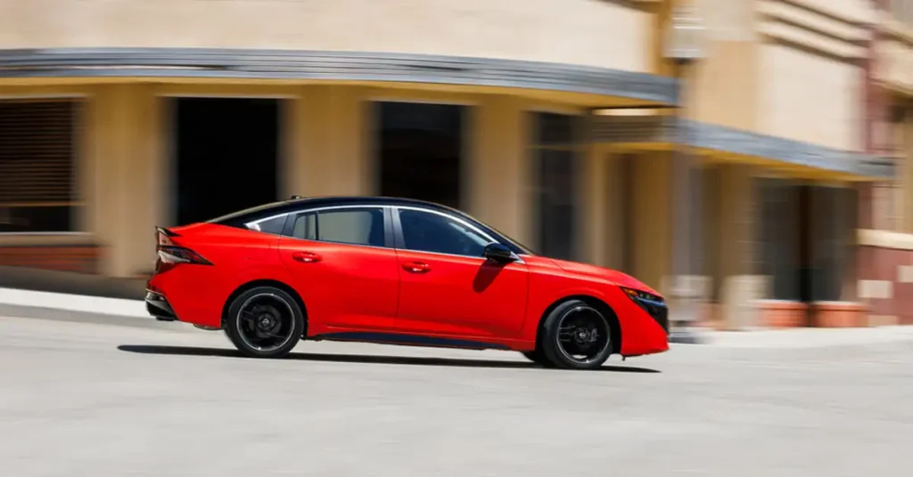 A vibrant, dynamic shot captures a red 2026 Nissan Sentra sedan in motion against an urban backdrop. The car, featuring a sleek black roof and black rims, is the focal point, its color popping against the neutral tones of the surrounding buildings.