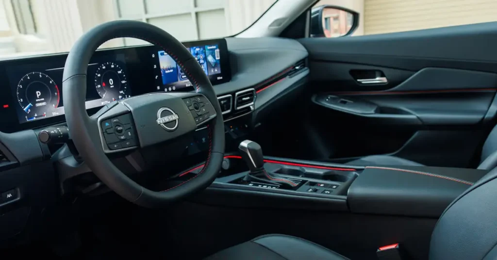 A detailed interior shot of a 2026 Nissan Sentra vehicle, focusing on the dashboard and front seating area. The interior is predominantly black with red accents, giving it a sporty and modern feel. The steering wheel, prominently displaying the Nissan logo, is positioned in the foreground, with a digital instrument cluster visible behind it.