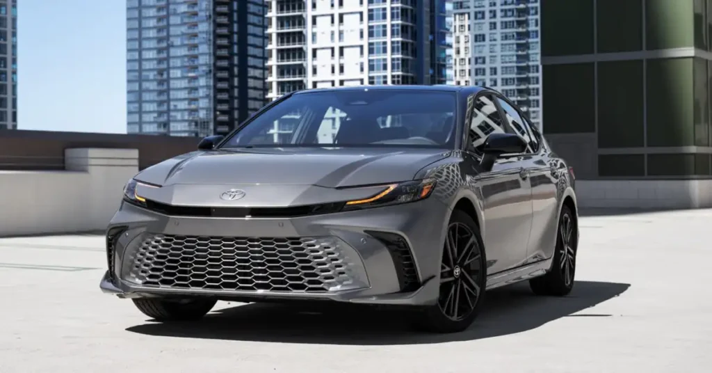 A gray 2026 Toyota Camry with black rims is parked on a concrete surface, with tall buildings in the background.