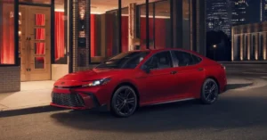 A red 2026 Toyota Camry parked on a city street at night, the car is facing left, the background includes a building with red curtains in the windows and a city skyline.