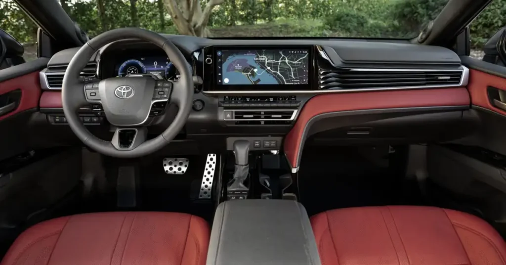 A 2026 Toyota Camry Nightshade car interior with a black dashboard and red leather seats, the steering wheel is gray with the Toyota logo in the center, the dashboard has a large screen displaying a map, the pedals are silver and have a sporty design, the center console is black with a gray armrest, the background shows trees and greenery, the overall style is modern and luxurious.