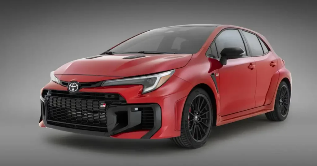 A red Toyota GR Corolla hatchback is displayed against a gray background. The car features a black grille with a "GR" badge, black wheels with red brake calipers, and black side mirrors. The car's design includes flared fenders and vents on the hood.