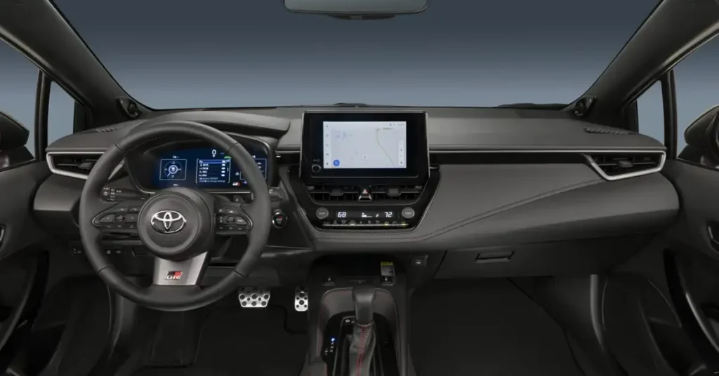 A detailed shot of the interior of a 2026 GR Corolla car, focusing on the dashboard and front console area. The color scheme is predominantly dark gray and black, giving it a sleek, modern appearance. The steering wheel, prominently displayed, features the Toyota logo at its center and is equipped with various control buttons. 