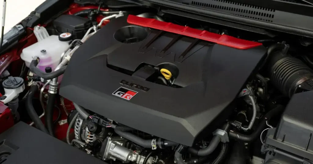 A close-up shot of a 2026 Toyota GR Corolla car engine bay, focusing on the engine itself. The engine is predominantly black with a red accent piece running across the top. The engine cover has the words "Turbo" and "GR" displayed on it.