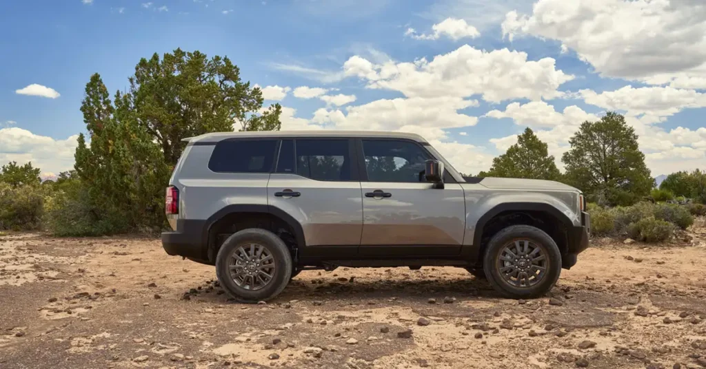 A silver The 2026 Land Cruiser SUV parked on a dirt road, with trees and a blue sky with white clouds in the background. The SUV is a four-door model with black trim around the wheel wells and along the bottom of the body.