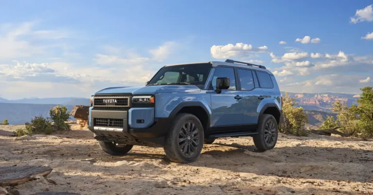 A light blue 2026 Toyota Land Cruiser SUV is parked on a rocky, sandy surface, with a backdrop of a blue sky dotted with fluffy white clouds and distant mountains. The SUV features a boxy design with a prominent front grille displaying the "TOYOTA" logo, black wheel arches, and matching black wheels. It has a roof rack and a two-tone color scheme with a lighter shade on the roof.