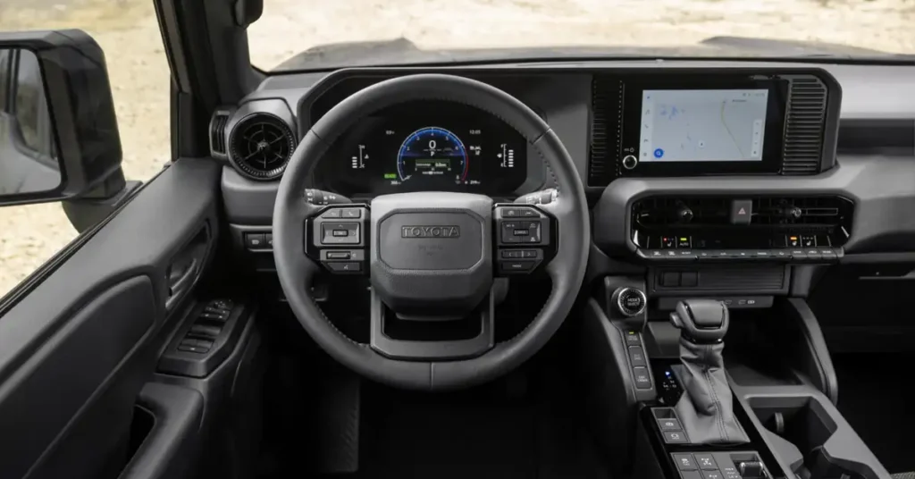 A digital photograph captures the interior of a 2026 Toyota Land Cruiser vehicle, focusing on the dashboard and steering wheel. The interior is predominantly black, with a modern and functional design. The steering wheel is centrally located, featuring the Toyota logo and various control buttons. 