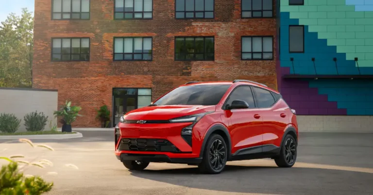 A vibrant red Chevrolet Bolt EUV is parked in front of a building with a mix of architectural styles. The car is positioned at a slight angle, showcasing its modern design and sleek lines.