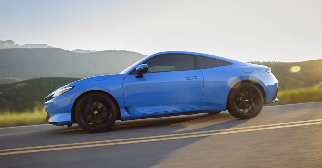 A light blue a 2025 Honda Prelude coupe car with black wheels is driving on a road with yellow lines, mountains in the background, and the sun shining in the distance.