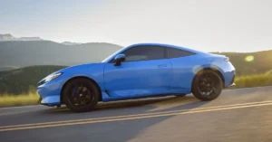 A light blue a 2025 Honda Prelude coupe car with black wheels is driving on a road with yellow lines, mountains in the background, and the sun shining in the distance.