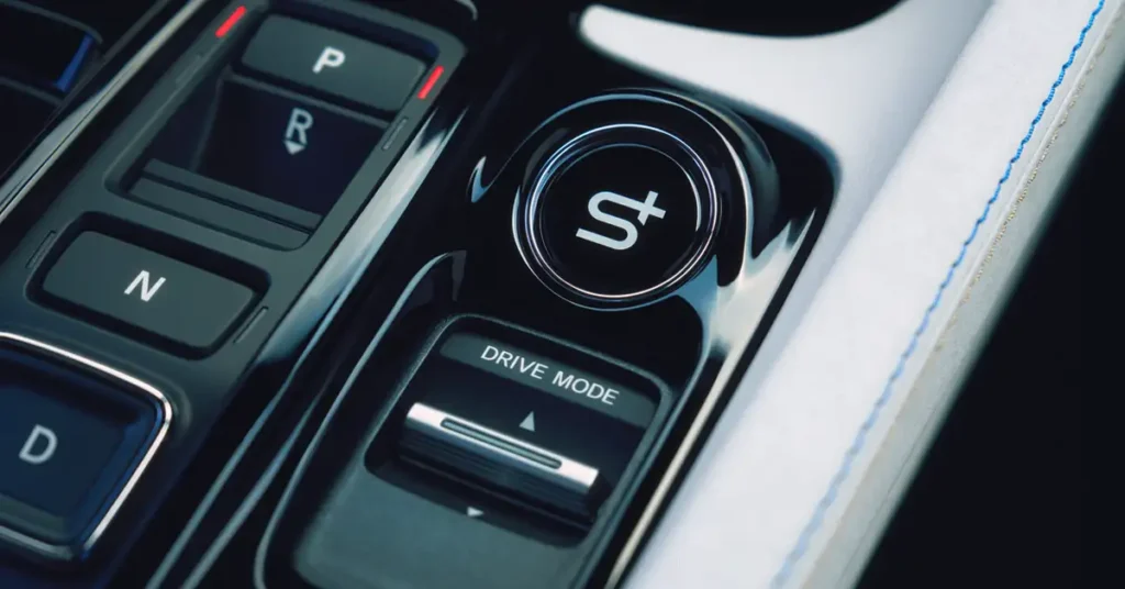 Close-up shot of a 2025 Honda Prelude center console, focusing on the gear shift area. The console is predominantly black with glossy finishes, featuring buttons labeled "P," "R," "N," and "D," each with white lettering. A larger, circular button with a stylized "S" symbol is positioned prominently, surrounded by a chrome ring. Below this, a rectangular button reads "DRIVE MODE" with an arrow symbol.