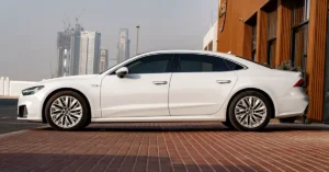 A side view of a white Audi A7 parked on a brick surface, with a modern building in the background. The car is sleek and sporty, with tinted windows and silver rims.