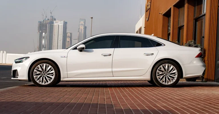 A side view of a white Audi A7 parked on a brick surface, with a modern building in the background. The car is sleek and sporty, with tinted windows and silver rims.