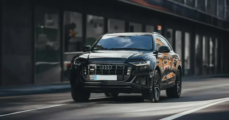 A black Audi Q8 SUV is captured in a low-angle, medium shot on an urban street. The car is in motion, with a slight blur indicating movement. The vehicle's front is prominently displayed, showcasing its grille, headlights, and the Audi logo.
