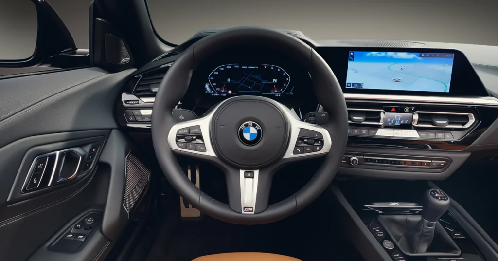 The interior of a BMW Z4 M40i , focusing on the dashboard and steering wheel. The steering wheel is black leather with silver accents and the BMW logo in the center. Behind the steering wheel is a digital instrument cluster displaying vehicle information.