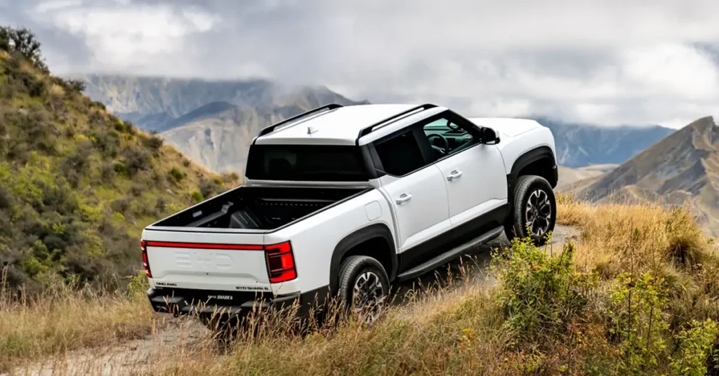A white BYD Shark 6 pickup truck is angled slightly to the right, ascending a hill covered in dry grass and sparse vegetation.