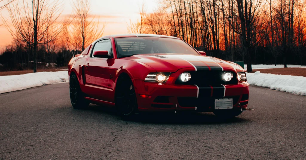 A low-angle, eye-level medium shot captures a red Ford Mustang with black racing stripes, parked on an asphalt road.