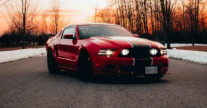 A low-angle, eye-level medium shot captures a red Ford Mustang with black racing stripes, parked on an asphalt road.
