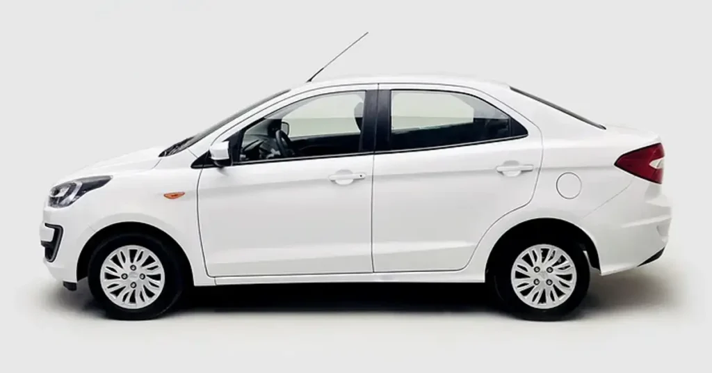 A side view of a white Ford Figo sedan car against a white background. The car has a sleek, modern design with a sloping roofline and a short rear deck. The wheels are silver with a multi-spoke design. The car is well-lit, and the white paint is clean and shiny.