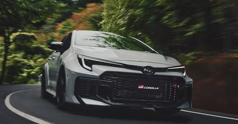 A gray Toyota GR Corolla is driving on a road with trees on either side. The car is in the foreground, and the background is blurred. The car is moving quickly, and the trees are a blur of green and brown. The car is a sporty hatchback with a black grille and black wheels.