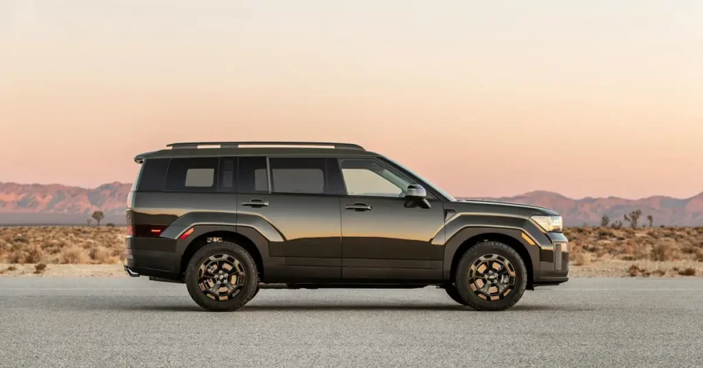 A side view of a dark gray Hyundai Santa Fe SUV parked on a paved road, with a desert landscape in the background under a soft, gradient sky. The SUV has a boxy design with a roof rack, tinted windows, and distinctive wheel rims. The desert background features sparse vegetation and distant mountains, all under a sky that transitions from a light orange near the horizon to a pale blue higher up.