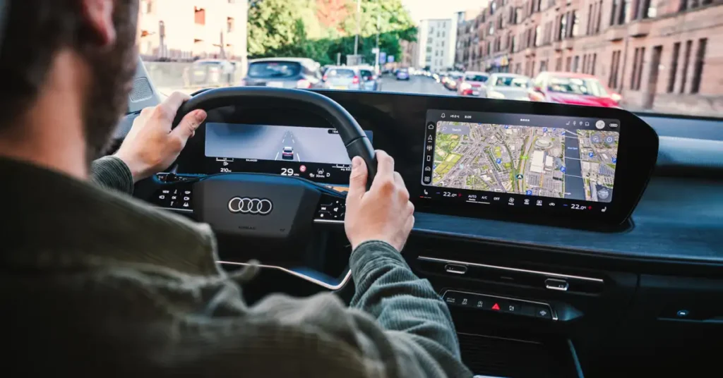 A medium shot captures the interior of 2025 New Audi Q3 car, focusing on a white male, around 30 years old, with light skin and brown hair, driving. His hands are on the steering wheel, and he is wearing a green corduroy shirt. The car's dashboard features a digital display showing a map and driving information. Outside the car, a city street is visible with buildings and other vehicles