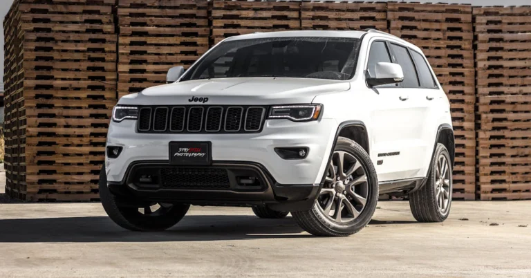 A front-angle shot of a white Jeep Grand Cherokee parked on a concrete surface, with a large stack of wooden pallets in the background.