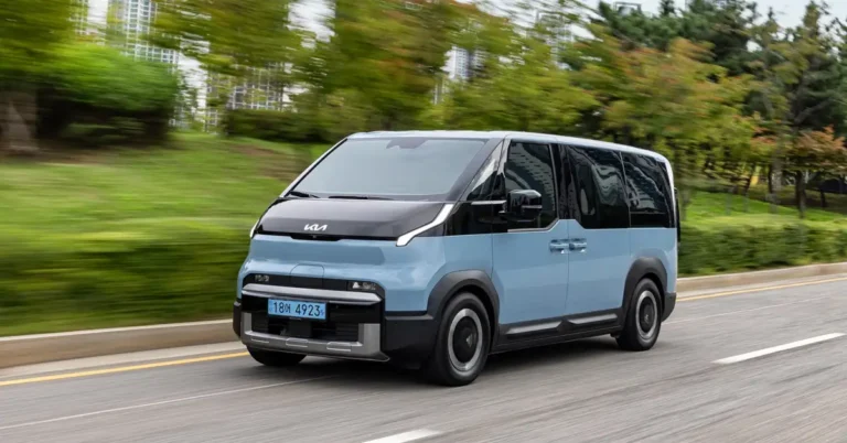 A light blue Kia PV5 van is driving on a road, with trees and buildings in the background. The van has a black hood and roof, and the Kia logo is visible on the front.