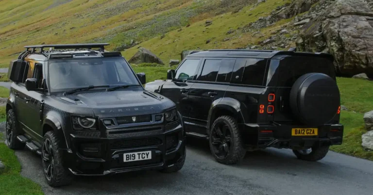 Two black Land Rover Defender SUVs are parked on a paved road, with a grassy hillside in the background. The SUV on the left has a roof rack and additional storage compartments on top.