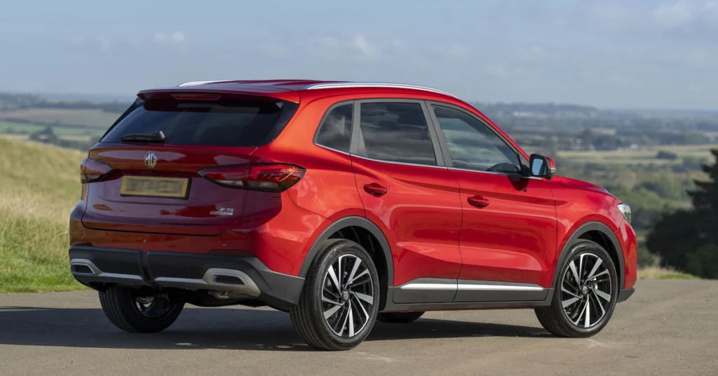 A vibrant, red MG MG ZS Hybrid+ SUV is captured from a low-angle, three-quarter rear perspective, set against a backdrop of rolling green hills under a clear blue sky.
