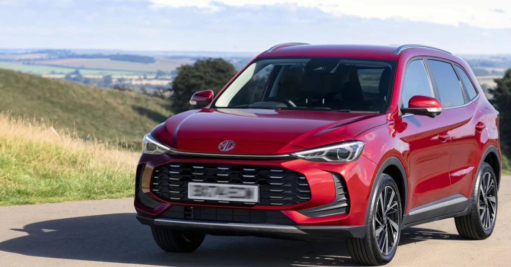 A vibrant, eye-level, medium shot captures a red MG ZS Hybrid SUV parked on a paved road, set against a backdrop of rolling green hills and a clear sky.