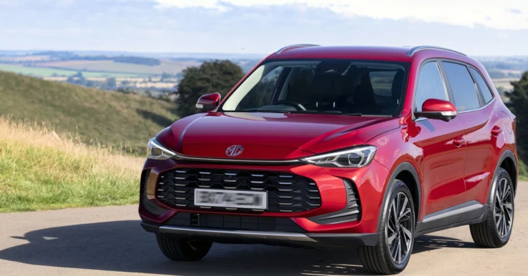 A vibrant, eye-level, medium shot captures a red MG ZS Hybrid SUV parked on a paved road, set against a backdrop of rolling green hills and a clear sky.