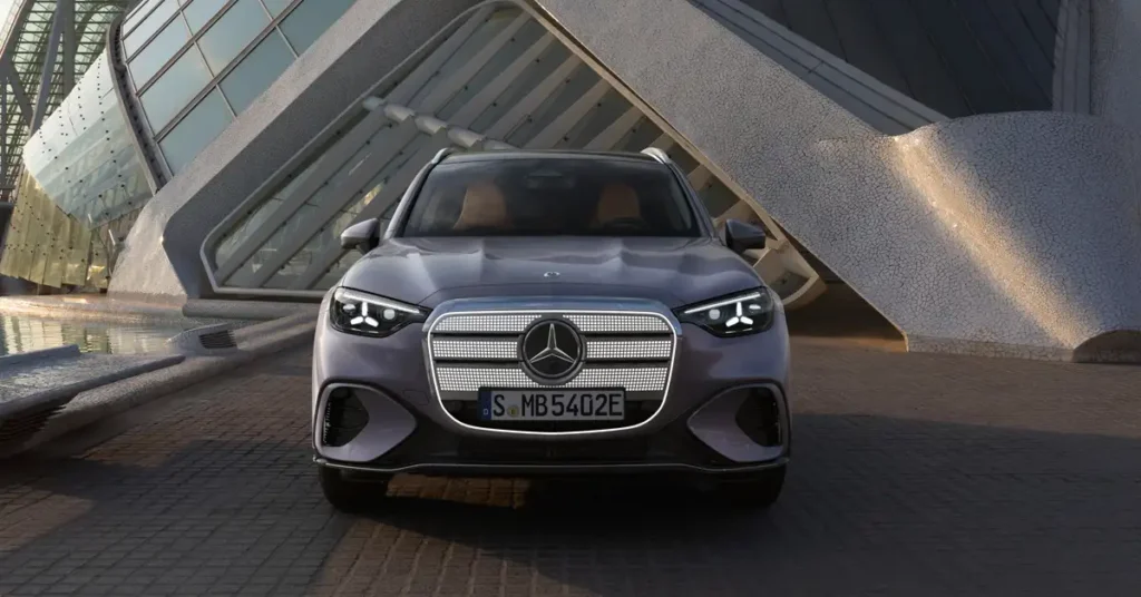 Front view of a gray Mercedes-Benz GLC EV 2026 with a silver grill and a license plate that reads "S MB 5402E" parked in front of a modern building with a gray and white exterior. The car is facing the camera and is positioned in the center of the frame.