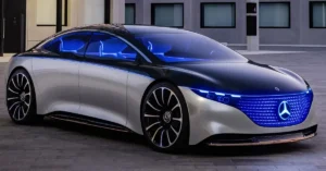 A Mercedes-Benz Vision EQS car with a sleek, aerodynamic design, featuring a two-tone color scheme with a silver body and a black roof. The car has a distinctive front grille illuminated with blue LED lights, and the headlights also have a blue hue.