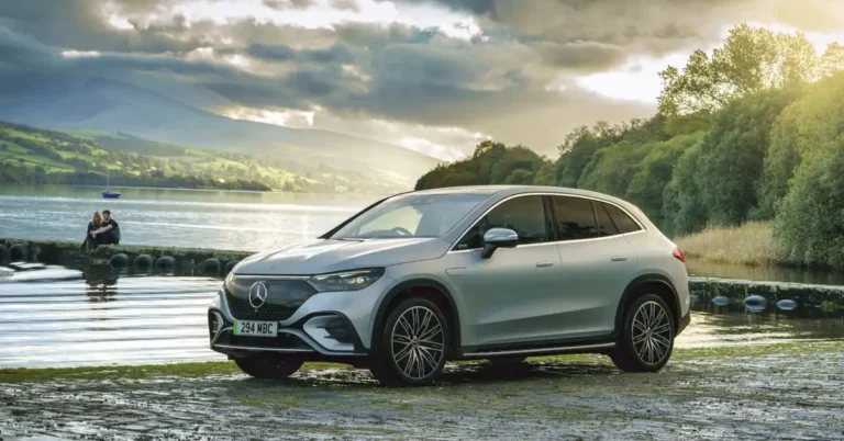 A silver Mercedes-Benz EQE SUV is parked on a muddy bank next to a lake. In the background, there are rolling green hills and a cloudy sky. A couple is sitting on a stone wall in the distance, looking out at the lake.