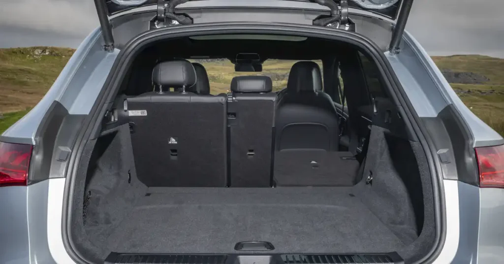 The open trunk of a gray Mercedes-Benz EQE SUV, showcasing a spacious interior with black carpeting and foldable rear seats, is captured in a well-lit, medium shot. The trunk's interior is clean and appears unused, with the rear seats partially folded down to extend the cargo area.