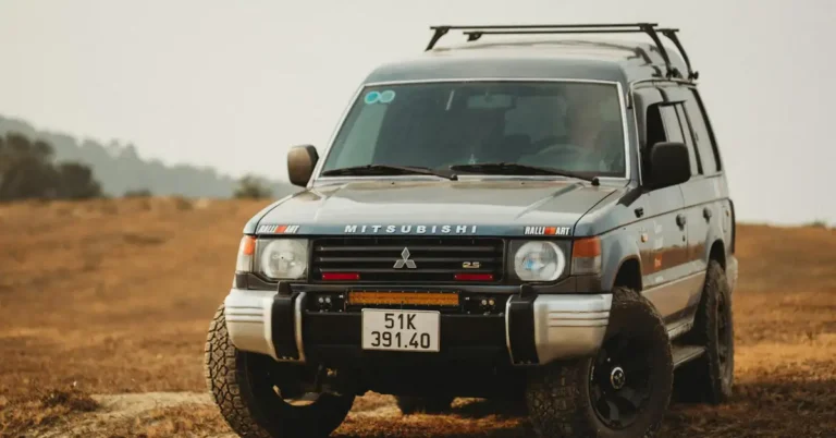A medium shot captures a Mitsubishi Pajero SUV, painted in a muted gray-blue, parked on a dirt surface. The vehicle is equipped with a roof rack and off-road tires, suggesting it is prepared for rugged terrain.