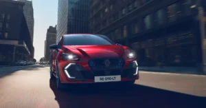 A red New Renault Clio 2025 full hybrid car is moving on a city street, the car is in the center of the image, the background shows tall buildings, the sky is clear and sunny, the image is a promotional shot.