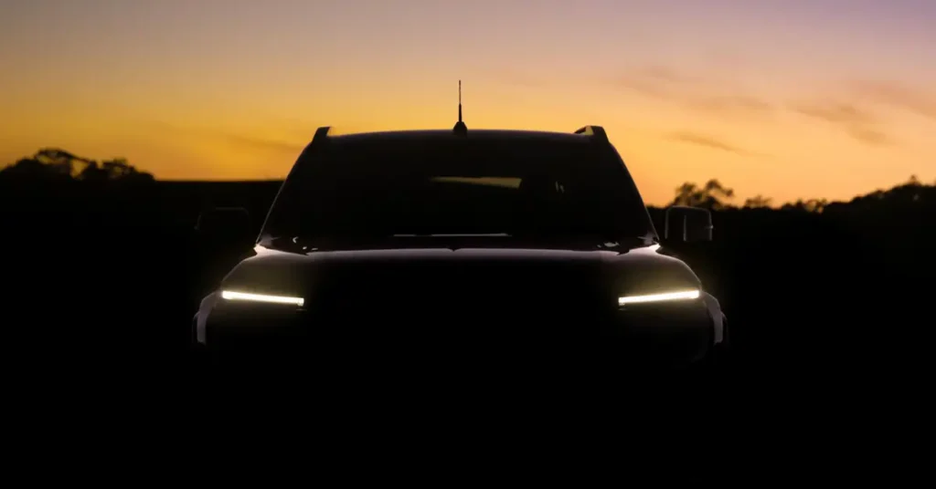 Front view of a black 2025 Nissan Navara SUV with headlights on, silhouetted against a sunset sky with orange and purple hues, the vehicle is parked on a dark field with trees in the background.