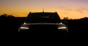Front view of a black 2025 Nissan Navara SUV with headlights on, silhouetted against a sunset sky with orange and purple hues, the vehicle is parked on a dark field with trees in the background.