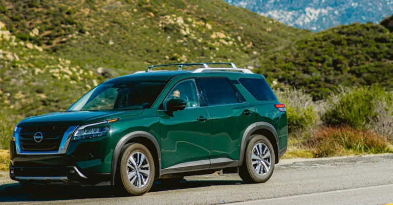 A side view of a dark green Nissan Pathfinder SUV parked on an asphalt road, with a backdrop of green hills and a mountain range in the distance. The SUV has a silver roof rack and silver rims on its tires.