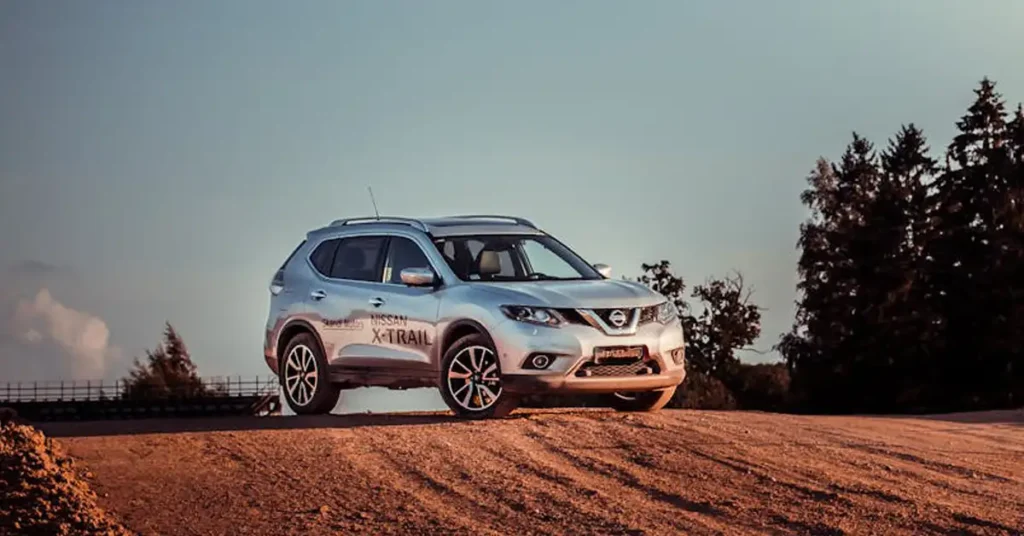 A silver Nissan X-Trail SUV is parked on a dirt hill, with tire tracks visible in the dirt. The sky is a pale blue, and there are trees in the background. The SUV is facing the viewer, and the Nissan logo is visible on the front