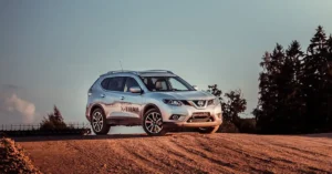 A silver Nissan X-Trail SUV is parked on a dirt hill, with tire tracks visible in the dirt. The sky is a pale blue, and there are trees in the background. The SUV is facing the viewer, and the Nissan logo is visible on the front
