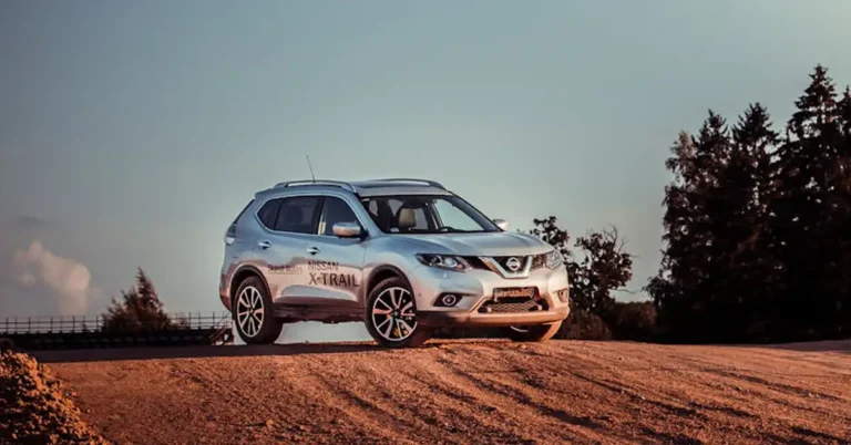 A silver Nissan X-Trail SUV is parked on a dirt hill, with tire tracks visible in the dirt. The sky is a pale blue, and there are trees in the background. The SUV is facing the viewer, and the Nissan logo is visible on the front